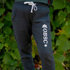A pair of gray OBSC Heritage Sweatpants with an adjustable drawstring waistband, displayed against a backdrop of greenery. The pants have white brand text and a graphic on the leg.