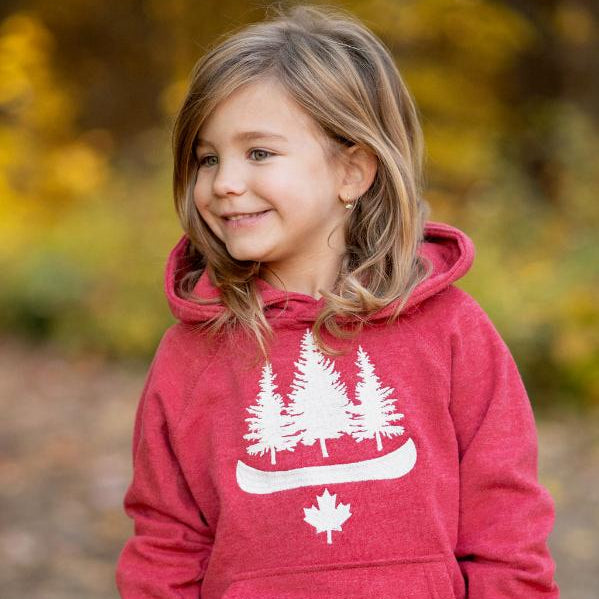 A child wearing a red hoodie with a white tree and Canadian maple leaf design, standing in a park with autumn leaves around.