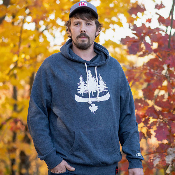 Adult male wearing a gray OBSC Heritage Hoodie with the logo visible on the front, standing outdoors with autumn leaves in the background.