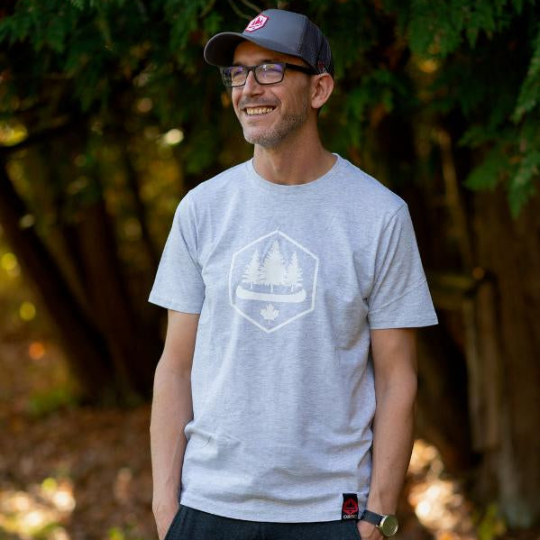 A man wearing a gray OBSC Heritage Logo Tee with the company's logo on the front, standing in a natural outdoor setting with trees in the background.